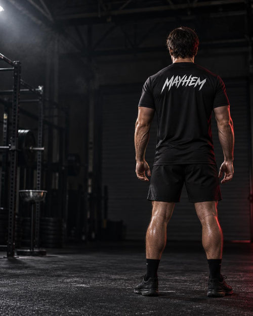 A man viewed from behind wearing a black athletic t-shirt with the word 'MAYHEM' in white stylized lettering across the back, paired with black shorts in a dark gym environment.