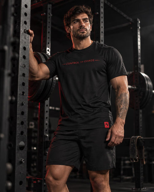 A muscular man in a gym wearing a black athletic t-shirt with red text reading 'CONTROL THE CHAOS' and matching black shorts, posing in front of a power rack.