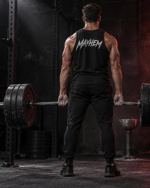 A muscular man viewed from behind in a gym, wearing a black tank top with the brand name 'MAYHEM' printed in white across the back, paired with black sweatpants and lifting a barbell.