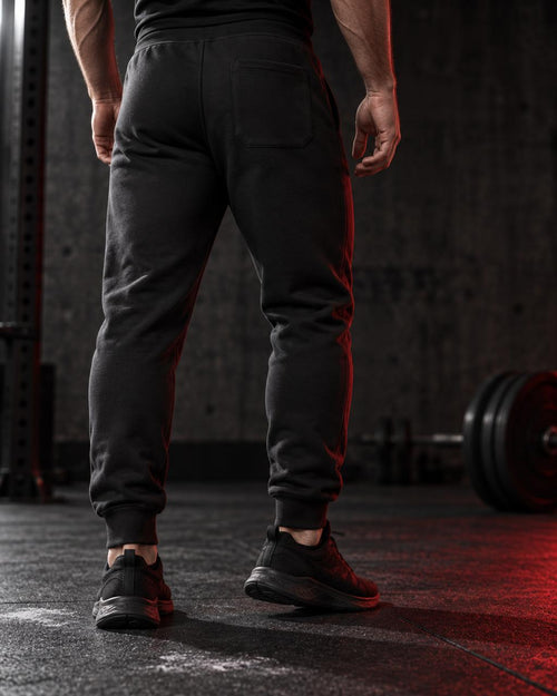 A person viewed from behind wearing dark charcoal gray jogger sweatpants with a subtle red side stripe, standing on a gym floor with barbell weights in the background.