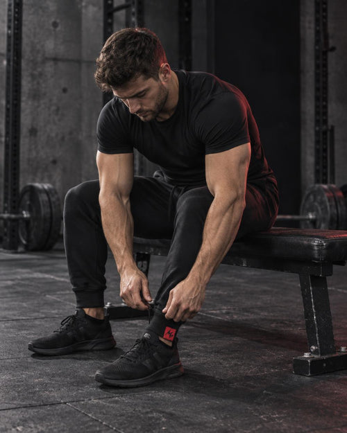 A man in a black athletic t-shirt and black joggers sits on a gym bench, adjusting the cuff of his black running shoe. The background is a dark, gritty gym environment with weight racks and concrete floors.