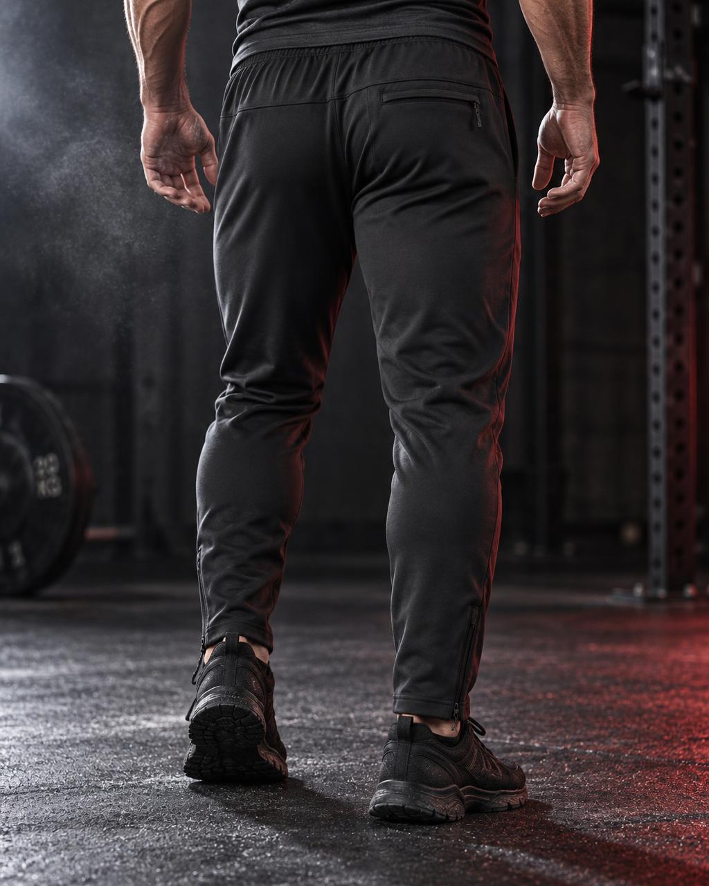 A person viewed from behind wearing black athletic jogger pants and black training shoes in a dark gym setting with red lighting.