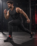 A man in a black athletic t-shirt and black jogger pants running through a dark gym, wearing black sneakers with a small red logo on the side.