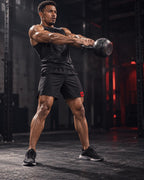 A muscular man in a gym setting, wearing a black tank top, black athletic shorts with a red logo, and black sneakers, holding a kettlebell in a front split stance.