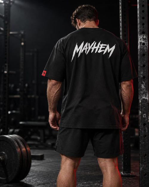 A man viewed from behind in a gym, wearing a black t-shirt with a large white 'MAYHEM' logo printed across the back and black athletic shorts.