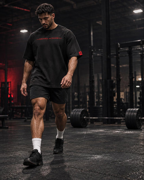 A man walking through a dark gym wearing a black short-sleeve t-shirt with red 'CORTEX ENERGY' text on the chest, black athletic shorts, white socks, and black sneakers.